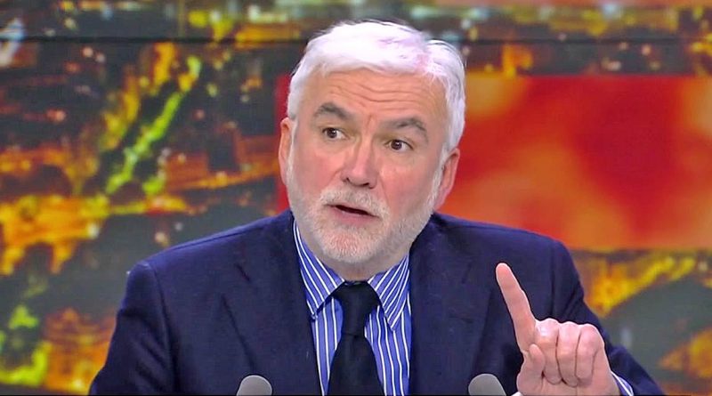 Homme aux cheveux gris et à la barbe blanche, en costume sombre et chemise à rayures, parlant en plateau TV et levant légèrement l’index devant un décor lumineux flou.