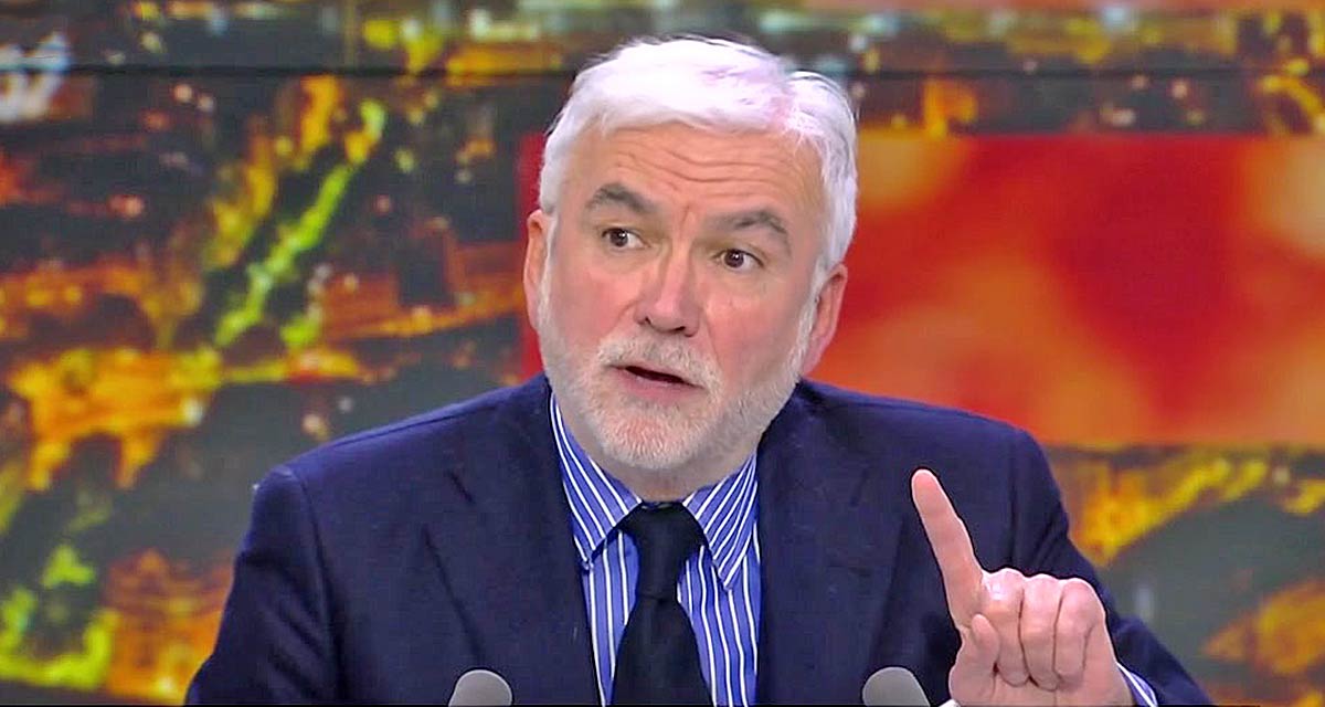 Homme aux cheveux gris et à la barbe blanche, en costume sombre et chemise à rayures, parlant en plateau TV et levant légèrement l’index devant un décor lumineux flou.