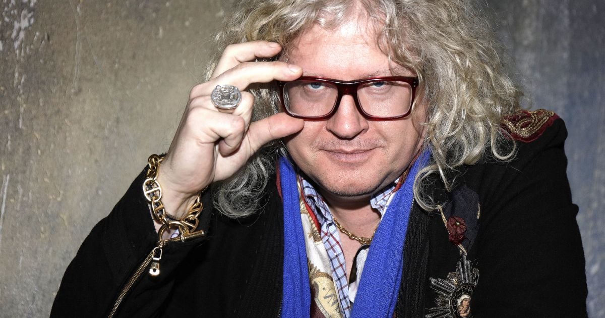 Homme aux cheveux blonds bouclés portant de grandes lunettes rectangulaires, posant face caméra avec un geste de la main près du visage, éclairage studio et fond neutre.