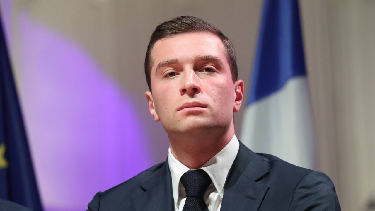 Homme en costume sombre et cravate, au visage sérieux, photographié devant un drapeau tricolore et un fond violet éclairé lors d’un événement officiel.