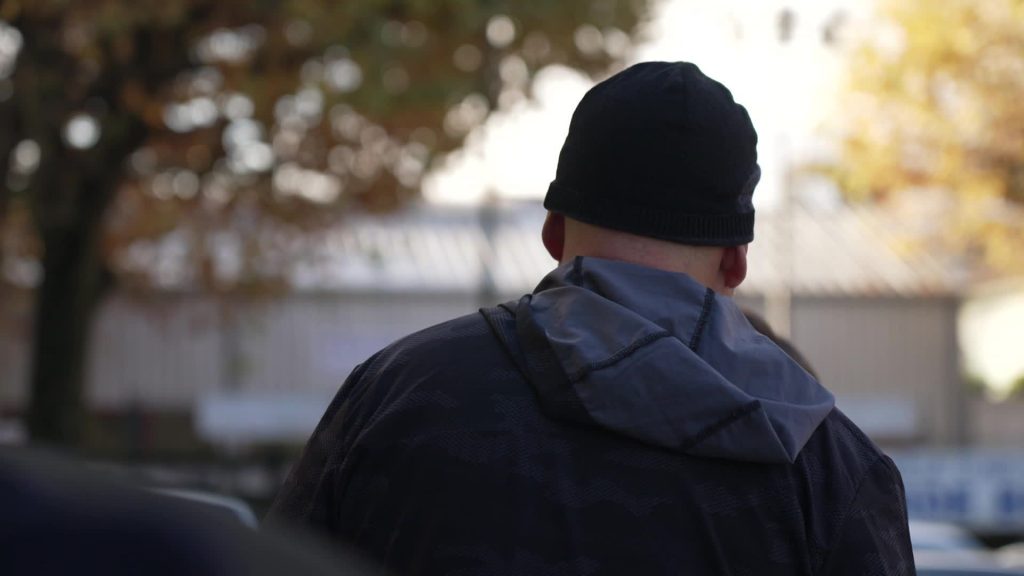 Un homme vu de dos, portant une veste sombre et un bonnet noir, se tient dehors par temps froid, symbolisant la solitude et la précarité d’un propriétaire contraint de vivre dans sa voiture après le squat de ses maisons.