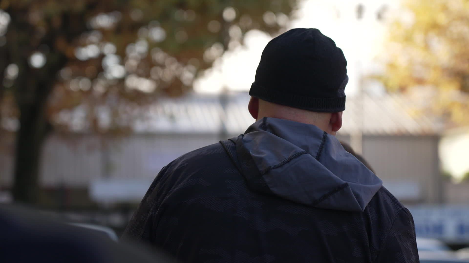 Un homme vu de dos, portant une veste sombre et un bonnet noir, se tient dehors par temps froid, symbolisant la solitude et la précarité d’un propriétaire contraint de vivre dans sa voiture après le squat de ses maisons.