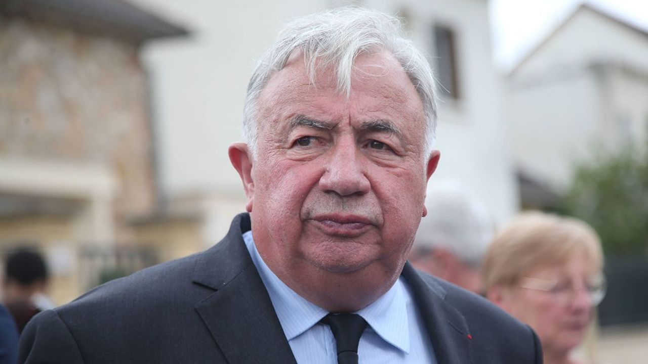 Homme âgé en costume sombre marchant en extérieur dans un cadre urbain flou, expression sérieuse.