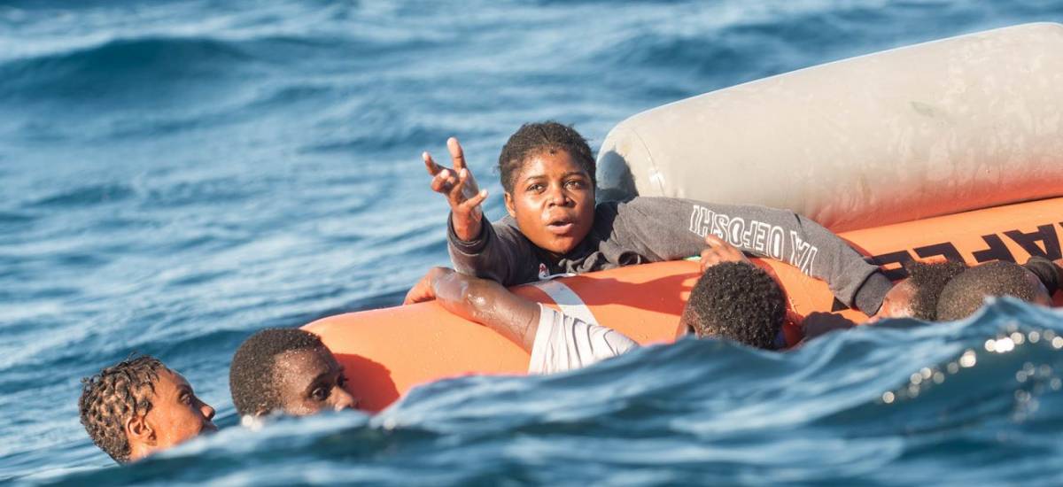 Personnes en détresse agrippées à un canot pneumatique en train de couler, au milieu de vagues, dont une femme tendant la main pour appeler à l’aide.