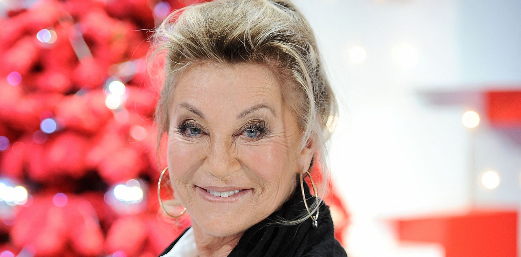 Gros plan d’une femme souriante aux yeux clairs, éclairée par une lumière douce, avec un fond flou rouge et blanc aux reflets lumineux.