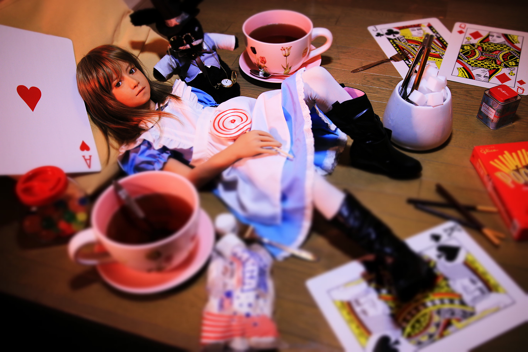 Poupée réaliste allongée au sol dans une mise en scène colorée, entourée de grandes cartes à jouer, de tasses de thé, d’objets décoratifs et de divers accessoires autour d’elle.