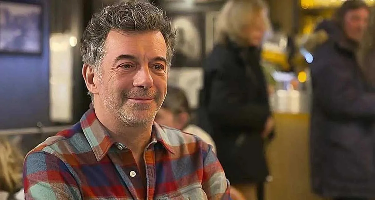 Homme souriant portant une chemise à carreaux, assis dans un café animé avec plusieurs personnes floues en arrière-plan.