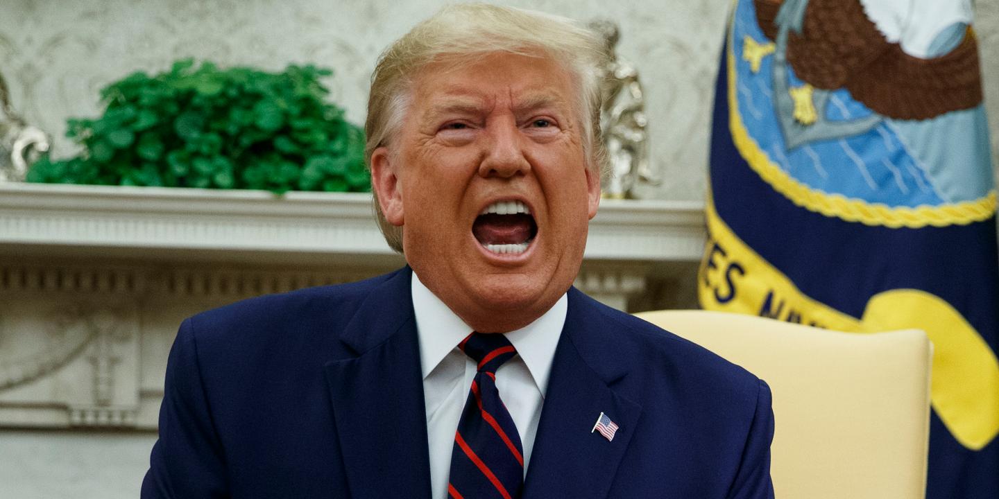 Homme âgé en costume bleu, visage rouge et bouche grande ouverte, apparaissant en pleine colère lors d’une prise de parole dans un bureau officiel orné de drapeaux et de décorations gouvernementales.