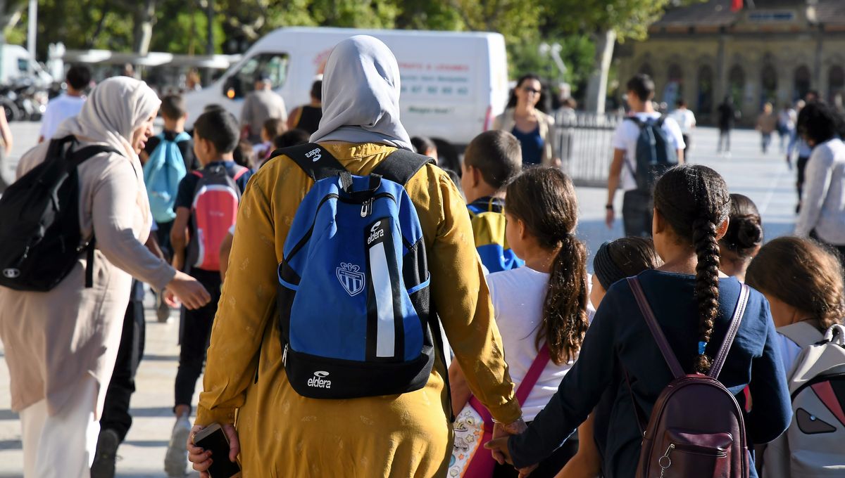 Groupe d’enfants marchant dans la rue, accompagnés par des adultes dont certaines portent un voile, scène de sortie d’école en milieu urbain.