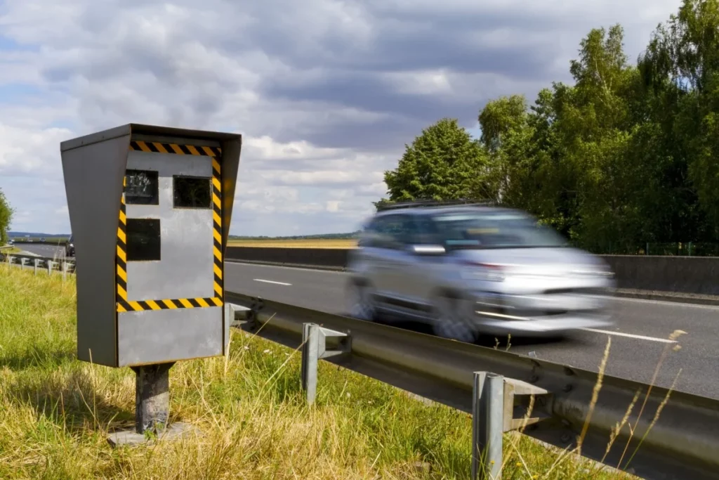 Radar automatique installé en bord de route avec un véhicule flou passant à vitesse modérée, environnement rural, ciel nuageux, scène illustrant la surveillance routière et le contrôle de vitesse