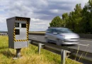 Radar automatique installé en bord de route avec un véhicule flou passant à vitesse modérée, environnement rural, ciel nuageux, scène illustrant la surveillance routière et le contrôle de vitesse