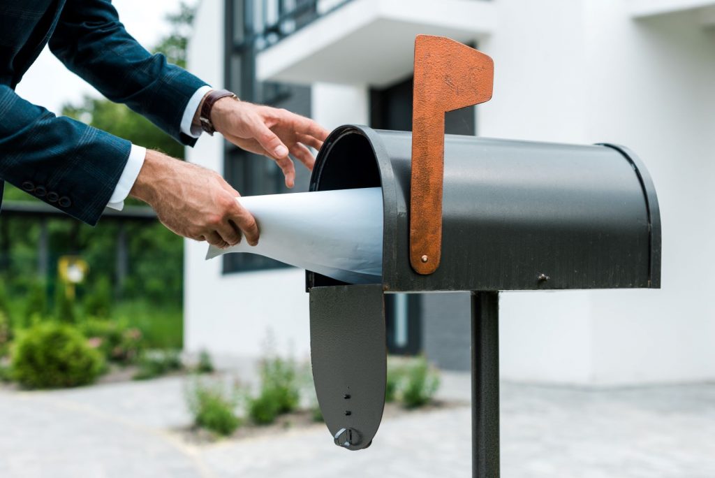 Personne glissant une grande enveloppe blanche dans une boîte aux lettres métallique en extérieur.