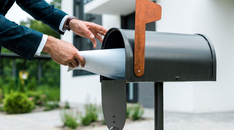Personne glissant une grande enveloppe blanche dans une boîte aux lettres métallique en extérieur.