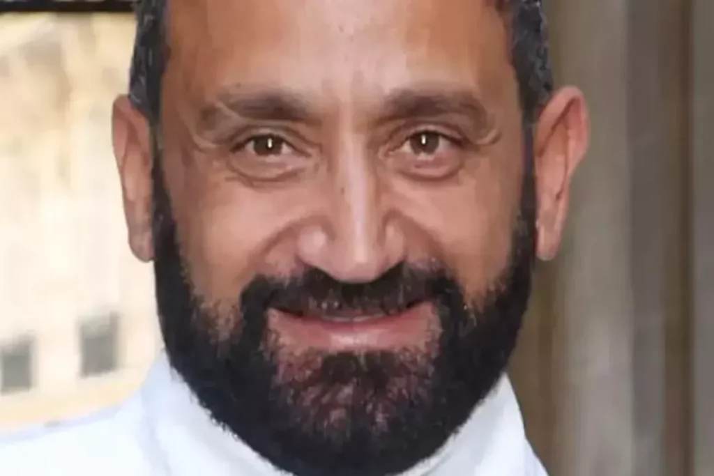 Homme barbu souriant regardant l’objectif, visage en gros plan, expression confiante, arrière-plan flou évoquant un cadre urbain
