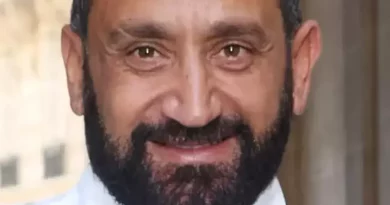 Homme barbu souriant regardant l’objectif, visage en gros plan, expression confiante, arrière-plan flou évoquant un cadre urbain