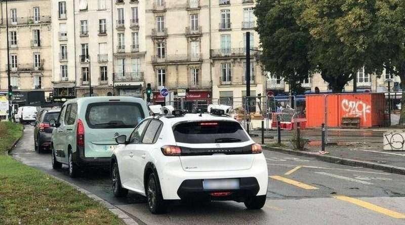 Voiture blanche équipée de caméras au toit roulant derrière un autre véhicule dans une rue urbaine un jour de pluie.