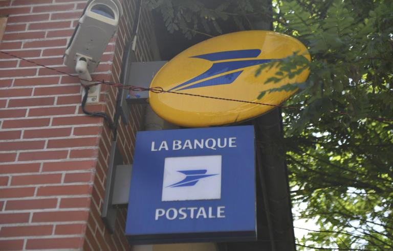 Enseigne bancaire bleue et jaune fixée sur la façade d’un bâtiment en briques, avec une caméra de surveillance et des arbres en arrière-plan, illustrant un établissement bancaire concerné par une panne informatique.