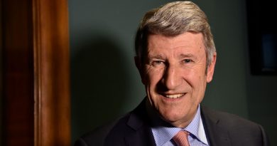 Homme mûr aux cheveux gris souriant face à l’objectif, portant un costume sombre et une cravate claire, dans un intérieur à l’éclairage doux