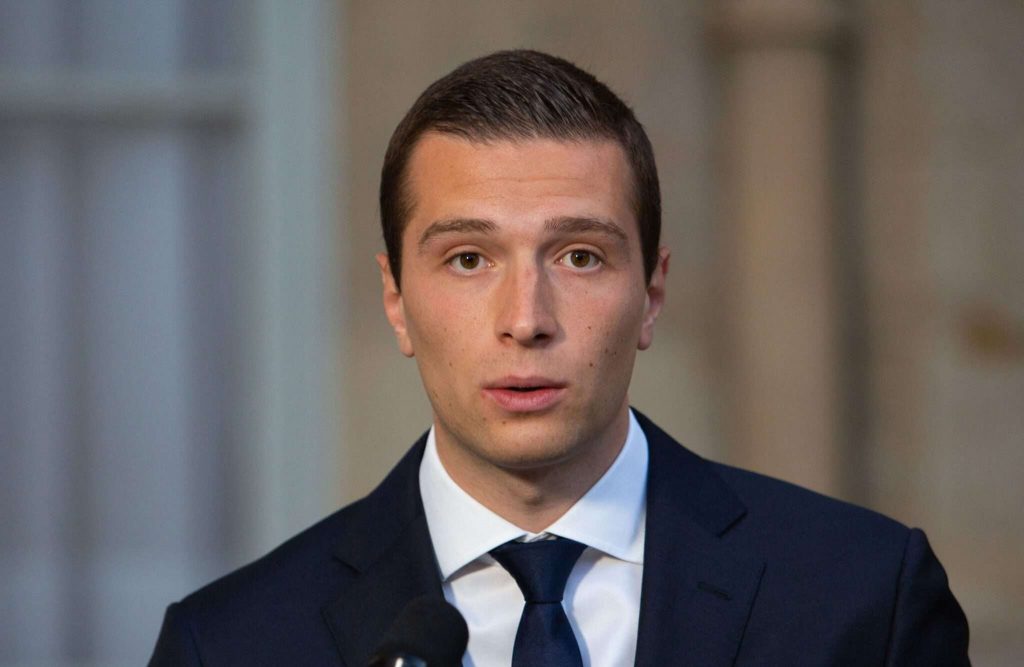 Jeune responsable politique en costume sombre, debout derrière un pupitre, regard sérieux face à l’objectif, dans un cadre institutionnel.