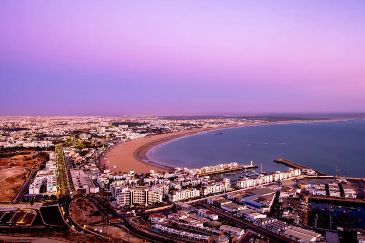 Vue aérienne d’une grande ville côtière au coucher du soleil, montrant une longue plage courbée, des immeubles modernes et une mer calme sous un ciel rose et violet.