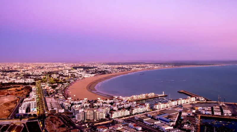 Vue aérienne d’une grande ville côtière au coucher du soleil, montrant une longue plage courbée, des immeubles modernes et une mer calme sous un ciel rose et violet.