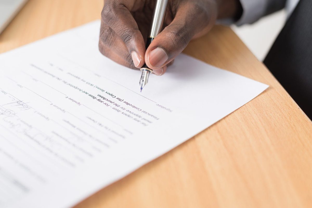Main tenant un stylo en train de signer un document professionnel posé sur un bureau en bois, illustrant une démarche administrative liée à un contrat de travail.