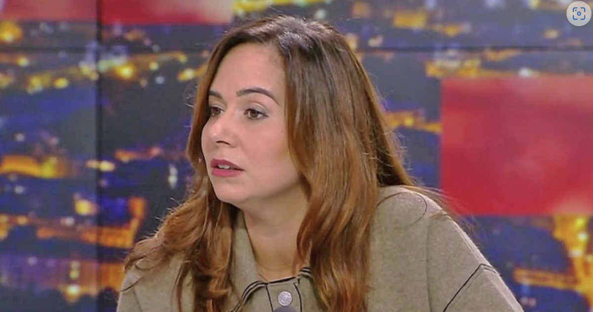 Femme assise sur un plateau de télévision, expression sérieuse, regard tourné vers un interlocuteur hors champ, ambiance de débat en direct sous éclairage de studio.