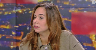 Femme assise sur un plateau de télévision, expression sérieuse, regard tourné vers un interlocuteur hors champ, ambiance de débat en direct sous éclairage de studio.