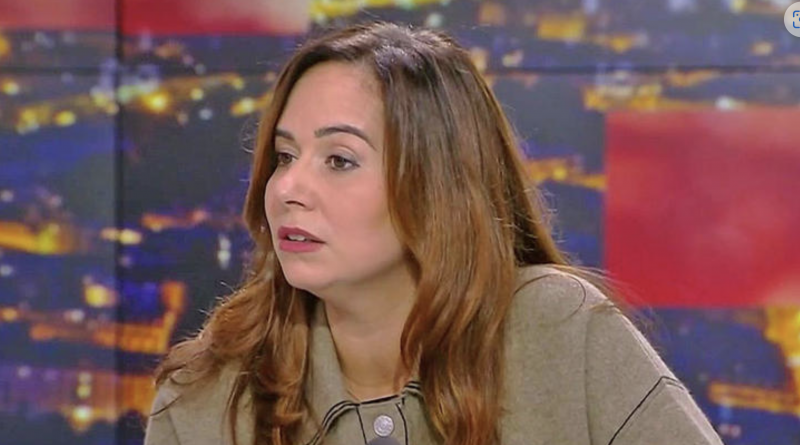 Femme assise sur un plateau de télévision, expression sérieuse, regard tourné vers un interlocuteur hors champ, ambiance de débat en direct sous éclairage de studio.