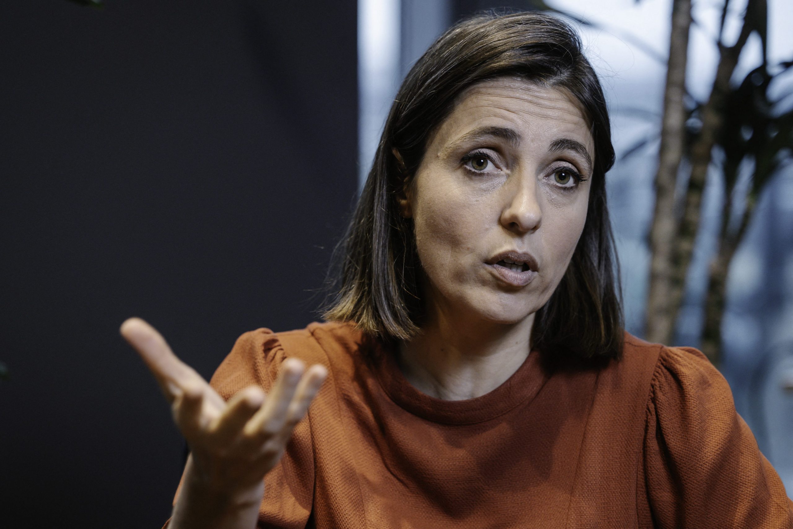 Femme parlant avec intensité, main levée pour appuyer son propos, assise en intérieur dans une atmosphère sérieuse.