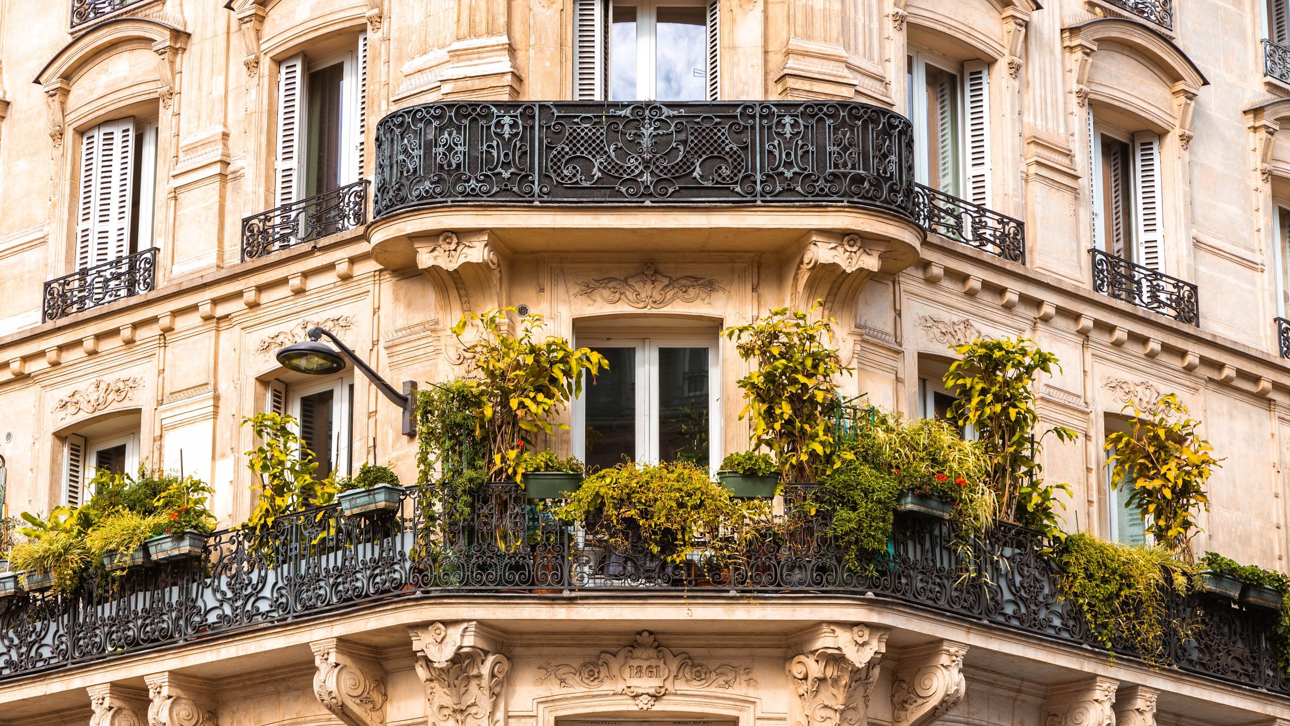 Façade d’un immeuble haussmannien parisien avec balcons en fer forgé et plantes luxuriantes, illustrant le charme mais aussi les coûts élevés liés à la propriété dans la capitale.