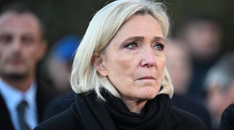 Personne au visage sérieux et ému, vêtue de noir, en extérieur, entourée d’autres individus en arrière-plan flou.