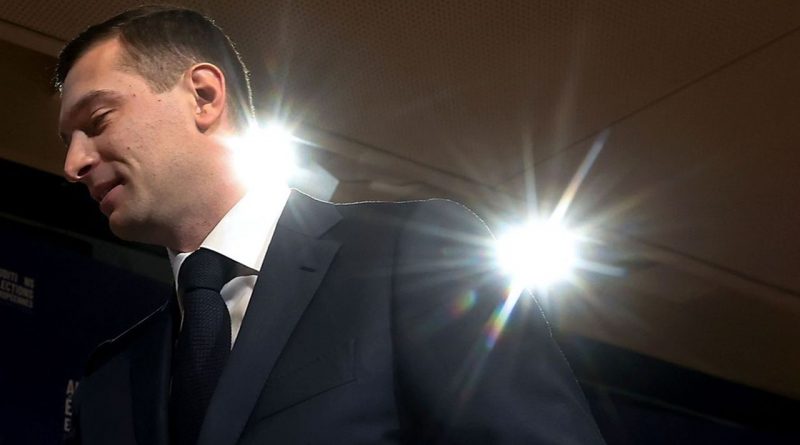 Un homme en costume photographié en contre-plongée sous un éclairage intense à l’intérieur d’un bâtiment.