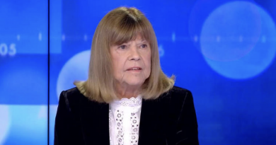 Femme âgée assise sur un plateau de télévision, visage sérieux et légèrement marqué par l’émotion, regard tourné vers un interlocuteur hors champ, fond bleu lumineux de studio.