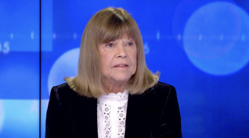 Femme âgée assise sur un plateau de télévision, visage sérieux et légèrement marqué par l’émotion, regard tourné vers un interlocuteur hors champ, fond bleu lumineux de studio.