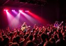 Concert rock sur une scène éclairée de lumières rouges et violettes, avec un chanteur et un guitariste face à une foule compacte de spectateurs levant les bras.