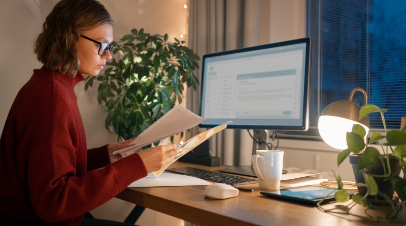 Personne assise à un bureau en train de consulter des documents devant un écran allumé, dans une pièce éclairée de manière chaleureuse.