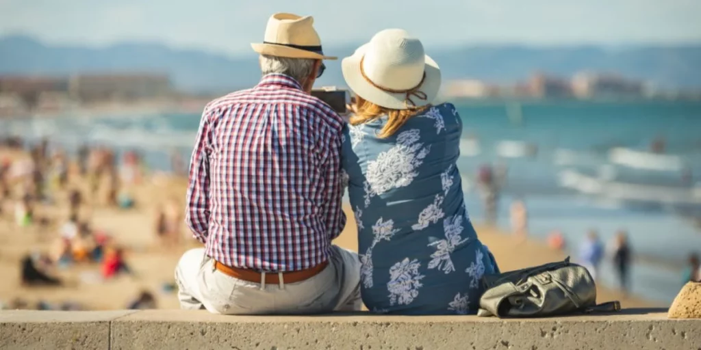 Couple de retraités assis face à la mer sur une promenade ensoleillée, profitant d’un cadre côtier paisible et d’un mode de vie détendu