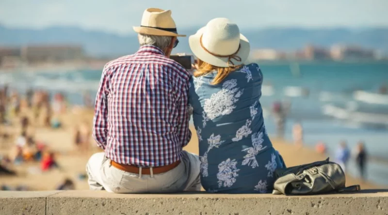 Couple de retraités assis face à la mer sur une promenade ensoleillée, profitant d’un cadre côtier paisible et d’un mode de vie détendu