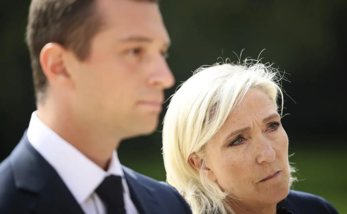 Portrait de deux personnes en tenue formelle, l’une au premier plan avec une expression sérieuse, l’autre légèrement floue à l’arrière-plan, en extérieur sous une lumière naturelle.