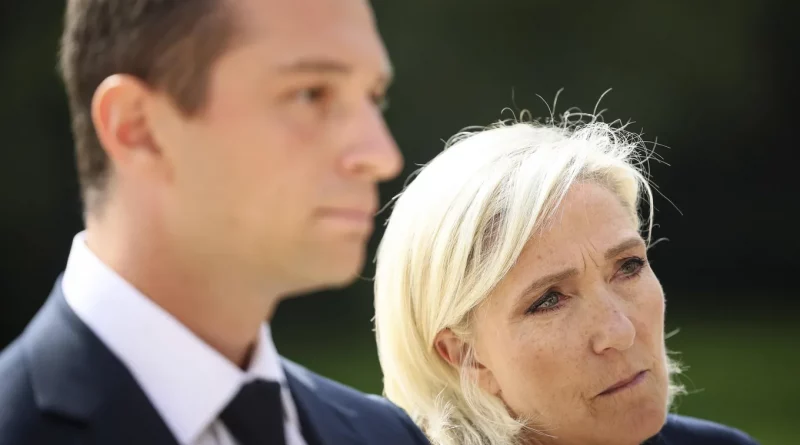 Portrait de deux personnes en tenue formelle, l’une au premier plan avec une expression sérieuse, l’autre légèrement floue à l’arrière-plan, en extérieur sous une lumière naturelle.