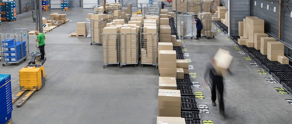 Des cartons empilés dans un grand entrepôt logistique avec des chariots métalliques et des employés en mouvement.