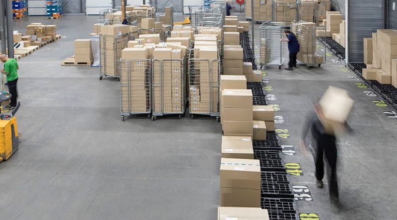 Des cartons empilés dans un grand entrepôt logistique avec des chariots métalliques et des employés en mouvement.