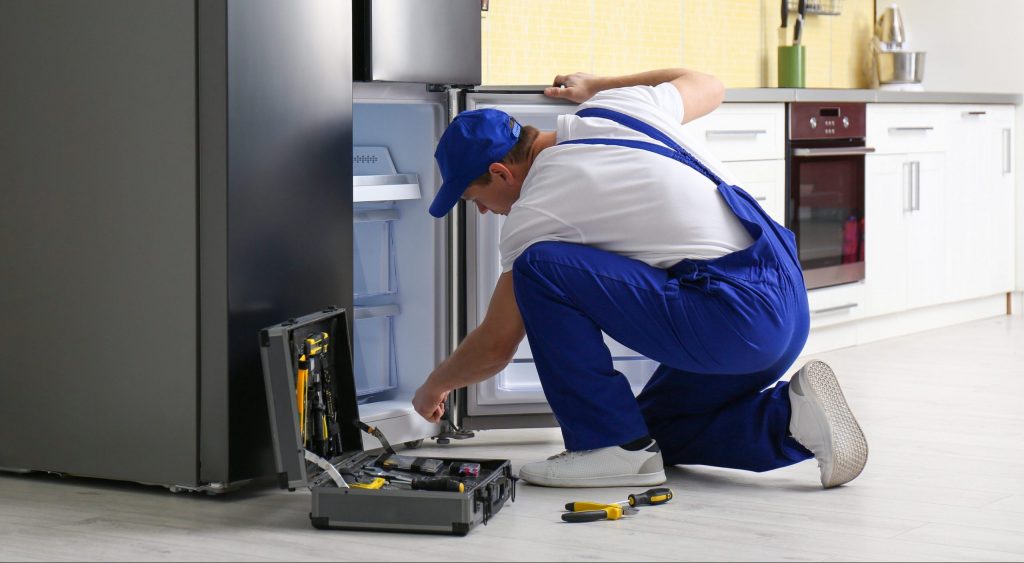Technicien réparant un réfrigérateur en panne dans la cuisine d’un logement meublé, outil ouvert au sol