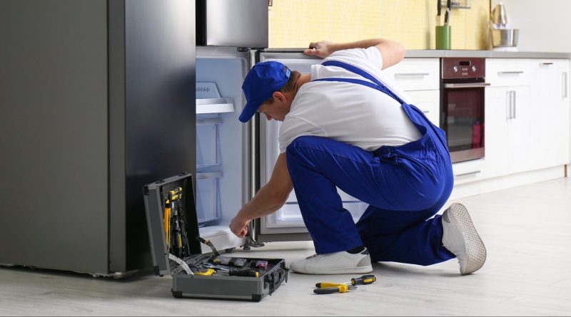 Technicien réparant un réfrigérateur en panne dans la cuisine d’un logement meublé, outil ouvert au sol