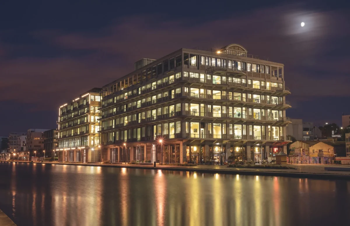 Façade d’un grand immeuble illuminé de nuit se reflétant sur l’eau, sous un ciel sombre éclairé par la lune.