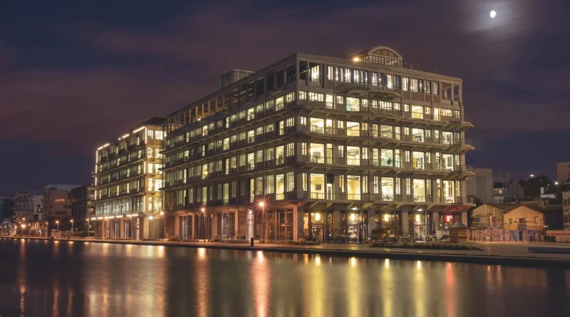 Façade d’un grand immeuble illuminé de nuit se reflétant sur l’eau, sous un ciel sombre éclairé par la lune.