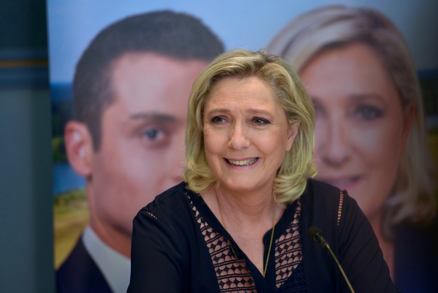 Portrait d’une femme blonde souriante assise devant un micro, avec deux visages flous en arrière-plan sur une affiche.