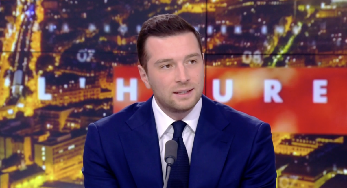 Personne en costume assise sur un plateau de télévision, parlant devant un micro, avec un décor lumineux en arrière-plan.