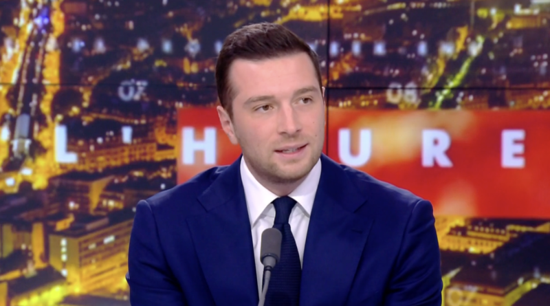 Personne en costume assise sur un plateau de télévision, parlant devant un micro, avec un décor lumineux en arrière-plan.